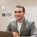 A person in a gray blazer sits at a table with a laptop, smiling slightly.