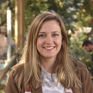 A person with long hair is smiling outdoors, with blurred greenery in the background.