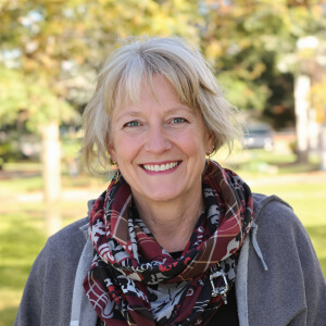 Smiling person with short blond hair wearing a patterned scarf and grey jacket, outdoors with blurred trees in the background.