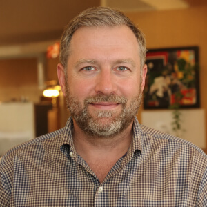A man with a beard and short hair smiles at the camera, wearing a checkered shirt. The background is an indoor setting with blurred decor.