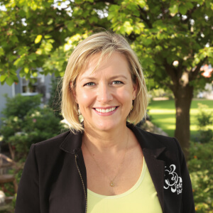 Woman with blonde hair smiling in an outdoor setting with green trees in the background, wearing a black jacket and yellow top.