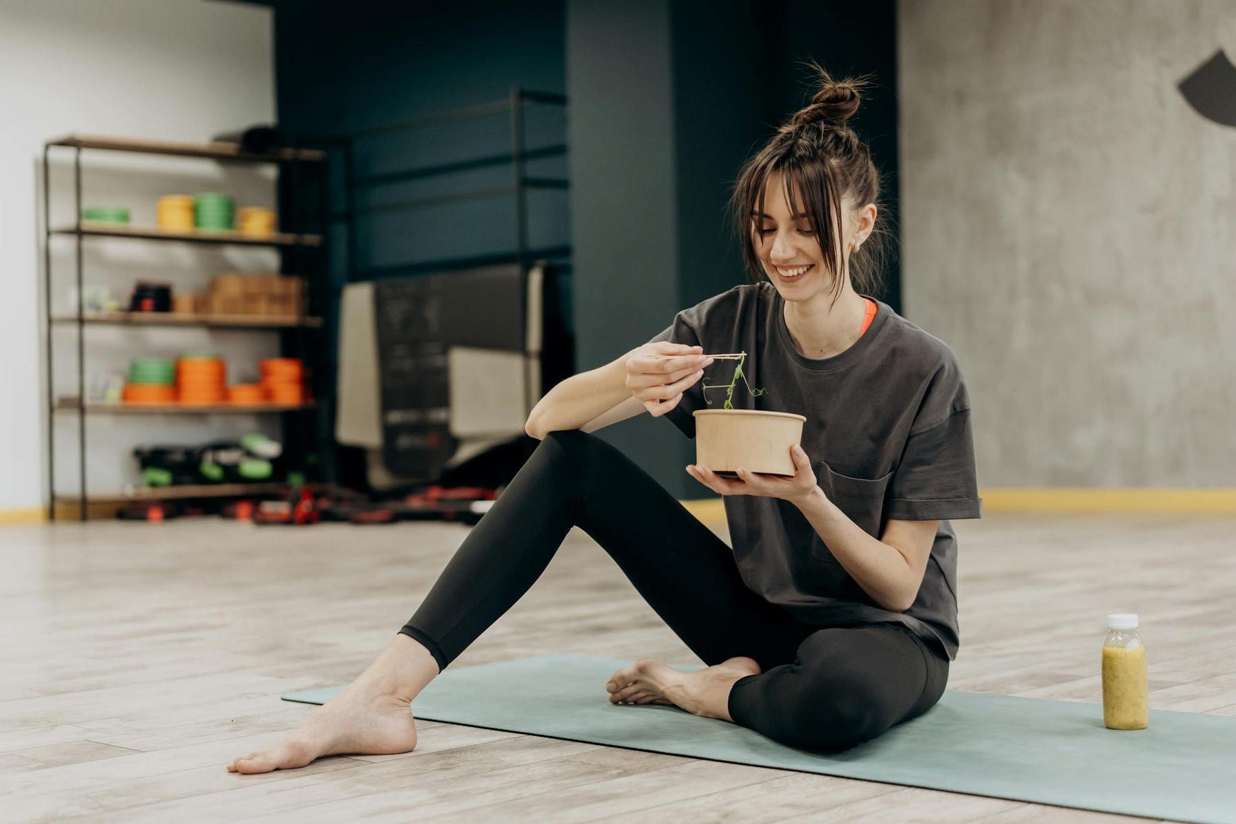 A woman sits cross-legged on a yoga mat indoors, eating from a bowl with chopsticks; a bottled drink is placed next to her.