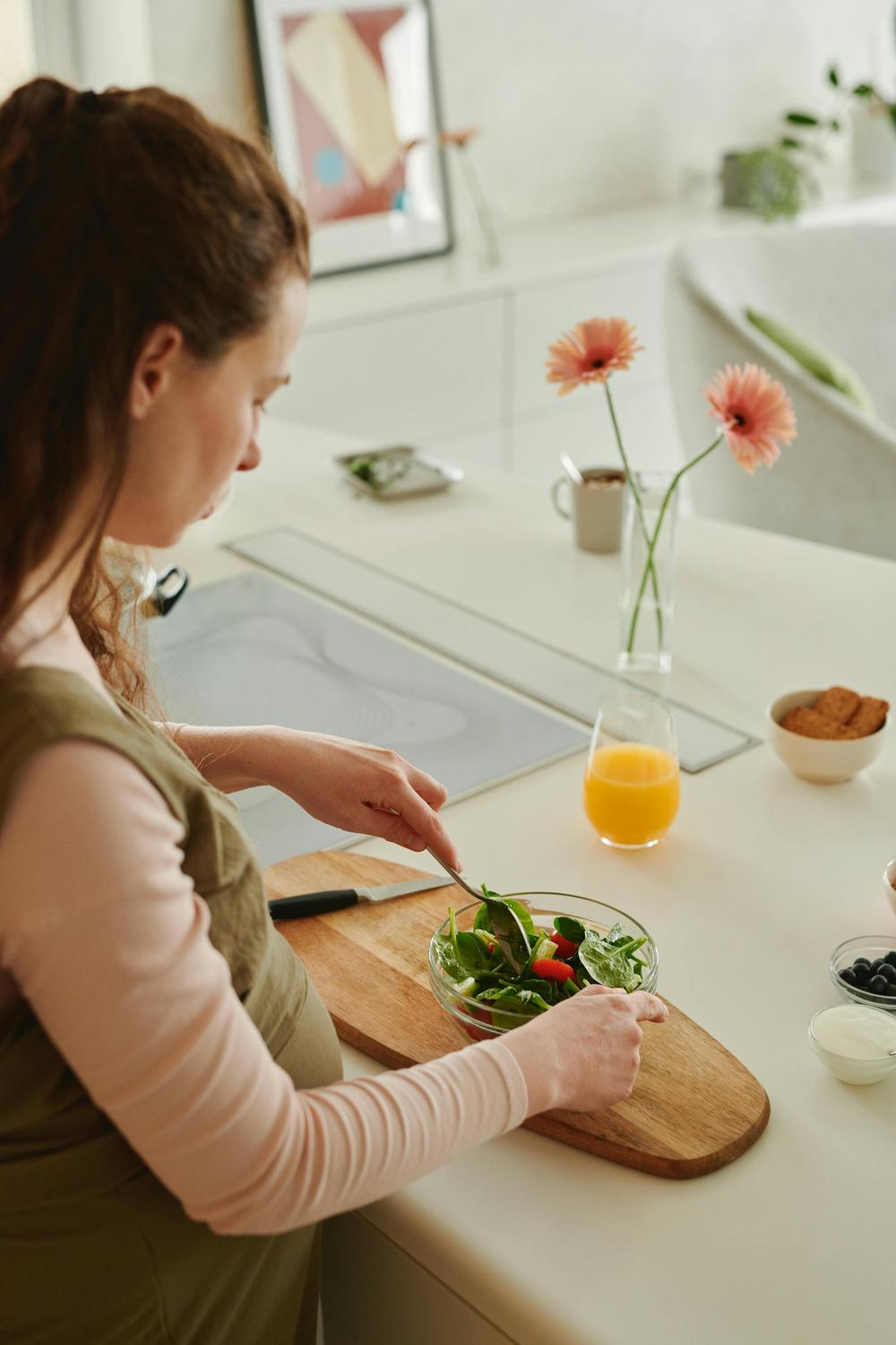 A person prepares a fresh salad in a glass bowl on a kitchen counter with a glass of orange juice, flowers, and small bowls of ingredients nearby.
