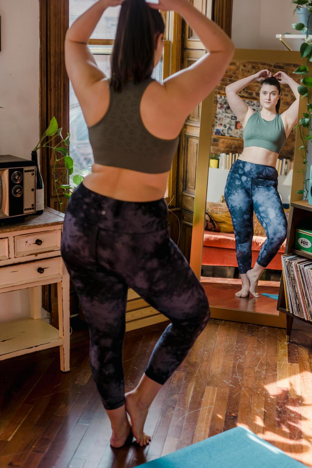 A person in athletic wear stands on a yoga mat and poses in front of a mirror in a sunlit room with wooden floors.