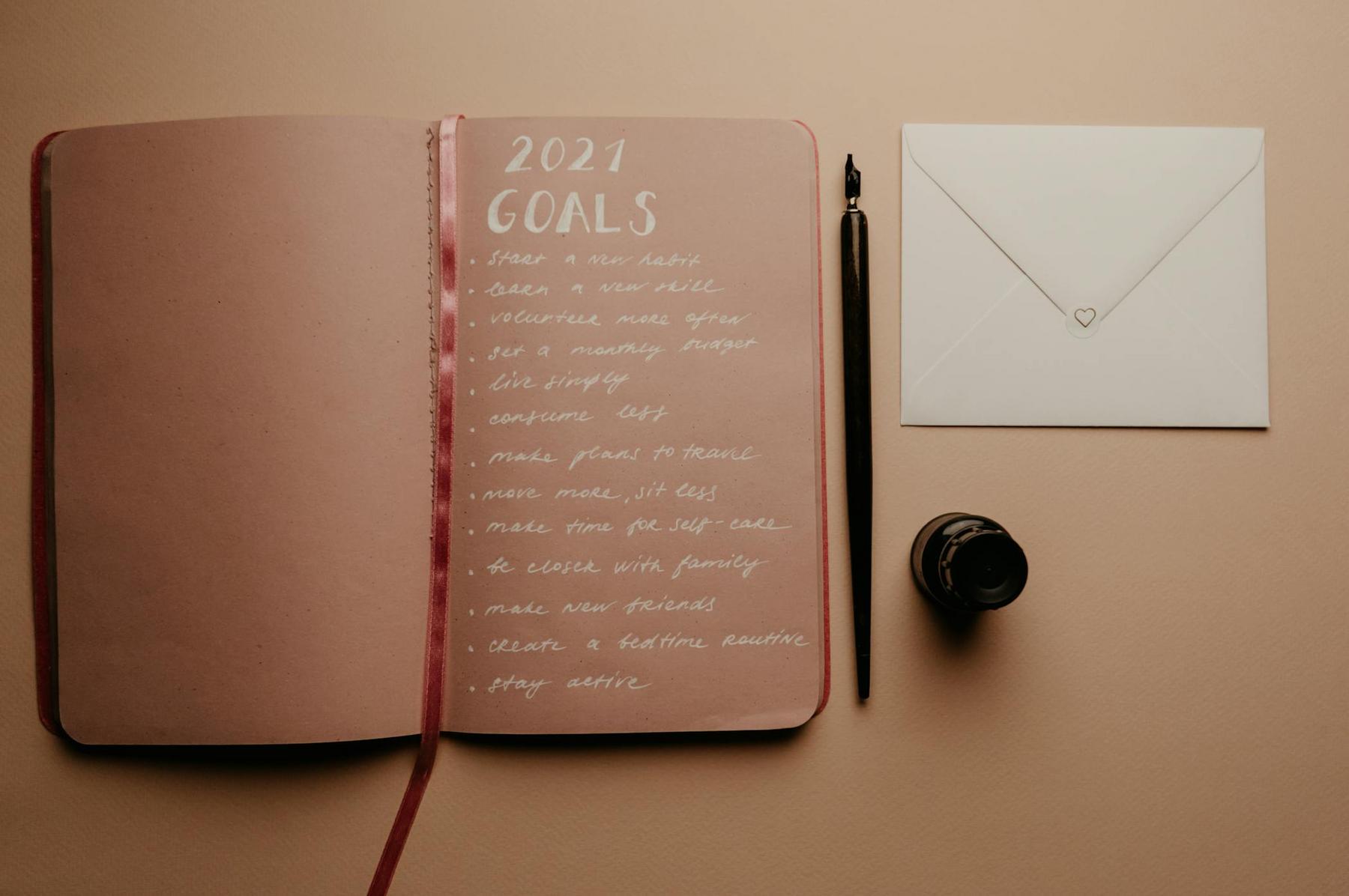 An open notebook lists 2021 goals in white ink, beside a black fountain pen, ink bottle, and a closed envelope on a beige surface.