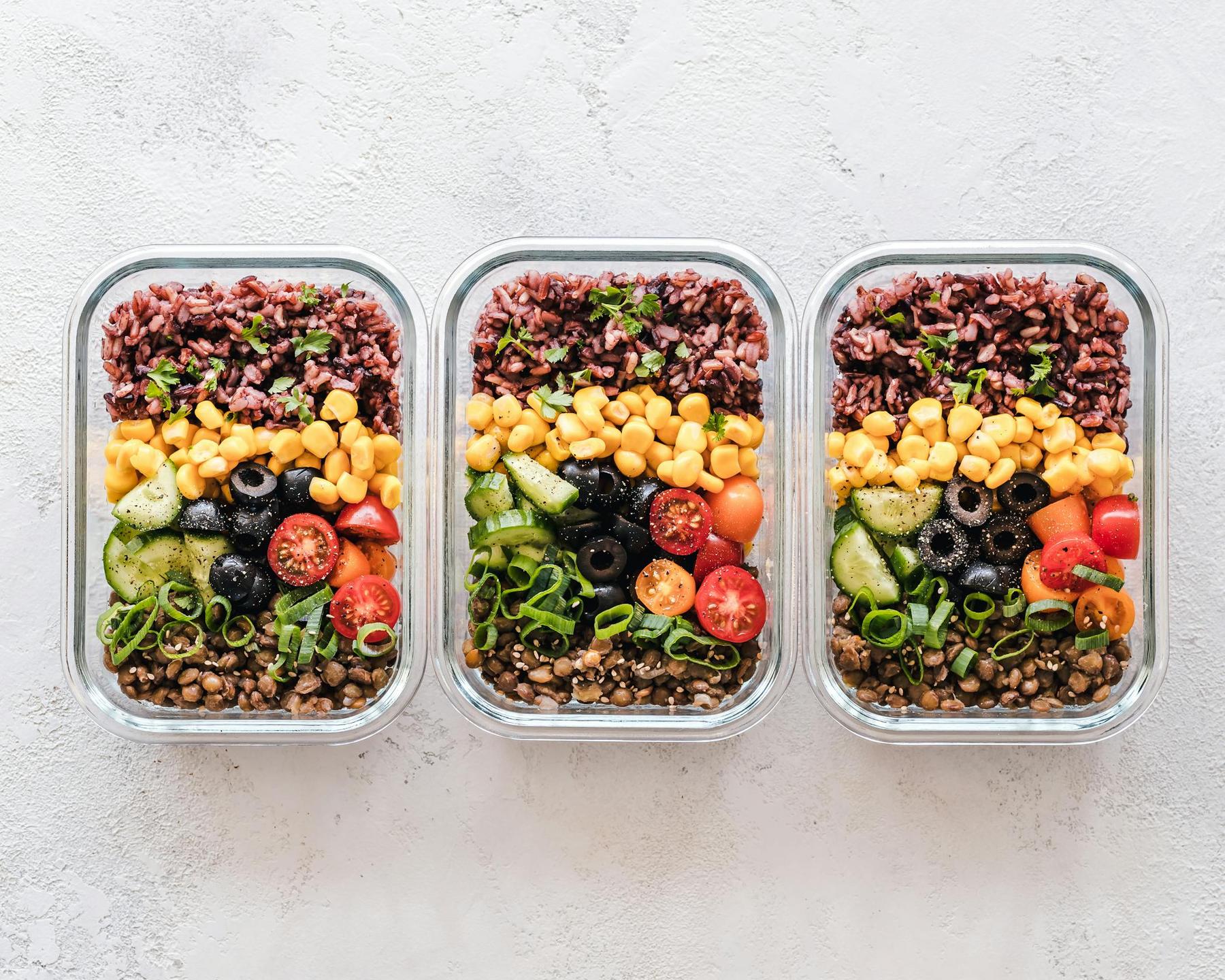Three glass meal prep containers filled with brown rice, sweetcorn, lentils, cherry tomatoes, cucumber, black olives, and chopped green onions, arranged in neat rows.