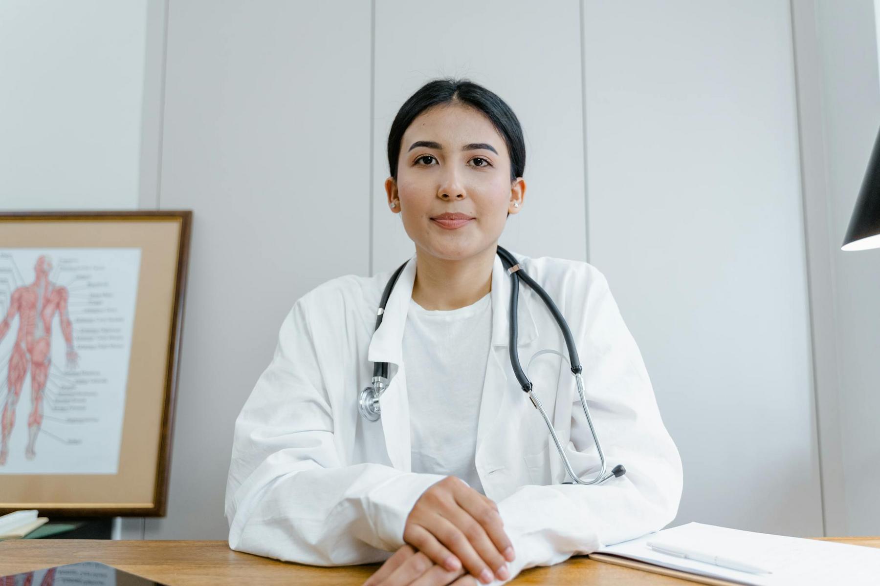 Doctor sitting at a desk with a stethoscope around her neck, anatomical poster in the background.