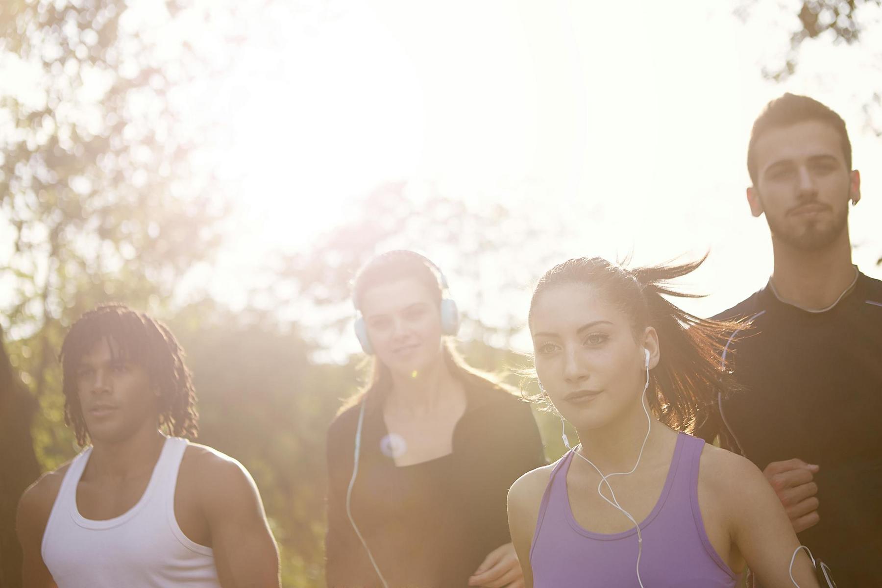 Four young adults jogging outdoors in sunlight, wearing athletic clothing and earphones, with trees blurred in the background.